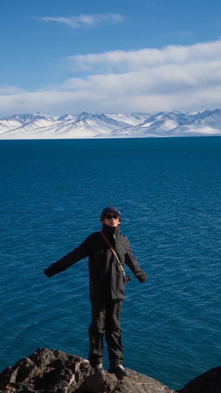 纳木错北岸，站在湖畔岩石上的人，身后是蓝色湖面与雪山。